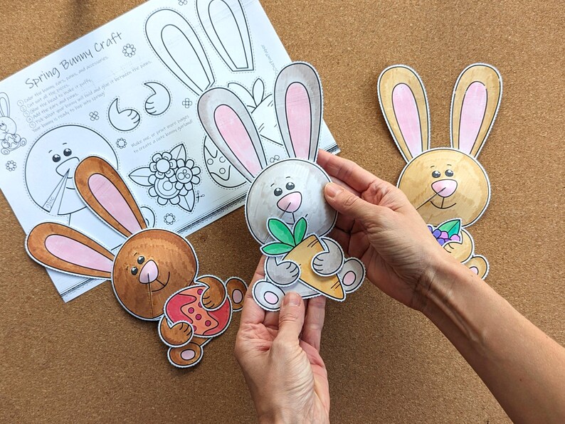 May include: A craft project featuring spring bunny cutouts. The bunnies are colored in shades of brown, gray, and tan, with pink inner ears. One bunny holds a carrot. A sheet of paper with instructions is also visible.