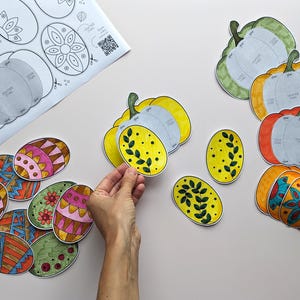 Op de afbeelding: Een verzameling papieren knutselwerkjes met kleurrijke, gedessineerde eieren en pompoenvormen. De set bevat een geprint instructieblad met diagrammen en diverse handgekleurde ovale en pompoenvormige papieren onderdelen met bloemmotieven.