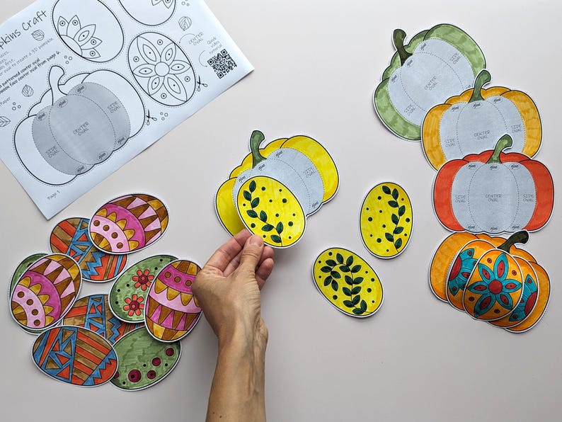 Op de afbeelding: Een verzameling papieren knutselwerkjes met kleurrijke, gedessineerde eieren en pompoenvormen. De set bevat een geprint instructieblad met diagrammen en diverse handgekleurde ovale en pompoenvormige papieren onderdelen met bloemmotieven.