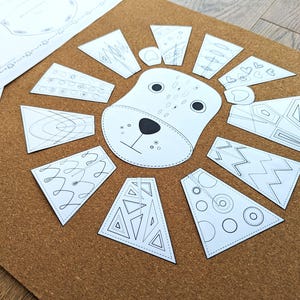 May include: A white lion face with black details and a paper mane made of various geometric shapes, arranged on a brown corkboard. The mane pieces have different patterns. A sheet of paper with a decorative border is in the upper left.