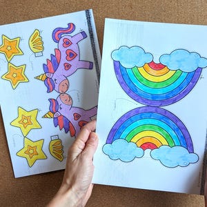 May include: Two sheets of paper with colourful illustrations. One sheet features a rainbow with blue clouds, and the other has unicorns, stars, and wings. The drawings are outlined in black and coloured with markers.