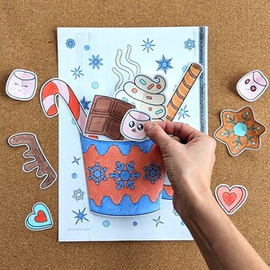 Peut inclure: Une main place un autocollant de guimauve dans un dessin d'une tasse de chocolat chaud. La tasse est d&eacute;cor&eacute;e de flocons de neige et remplie de cr&egrave;me fouett&eacute;e, de chocolat et d'un sucre d'orge. D'autres autocollants incluent un biscuit en pain d'&eacute;pices et un c&oelig;ur.
