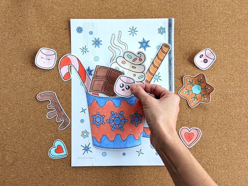 Peut inclure: Une main place un autocollant de guimauve dans un dessin d'une tasse de chocolat chaud. La tasse est d&eacute;cor&eacute;e de flocons de neige et remplie de cr&egrave;me fouett&eacute;e, de chocolat et d'un sucre d'orge. D'autres autocollants incluent un biscuit en pain d'&eacute;pices et un c&oelig;ur.