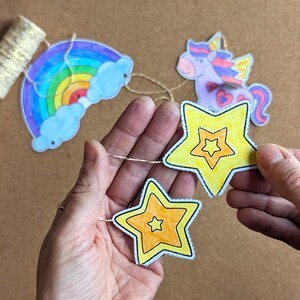 May include: A colourful garland featuring a rainbow, a unicorn, and yellow stars. The rainbow has blue clouds and the unicorn has wings. The stars are yellow with orange centres. The garland is strung on a thin, natural-coloured twine.