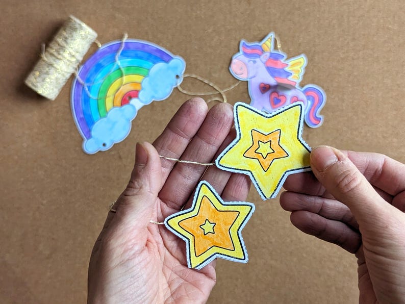May include: A colourful garland featuring a rainbow, a unicorn, and yellow stars. The rainbow has blue clouds and the unicorn has wings. The stars are yellow with orange centres. The garland is strung on a thin, natural-coloured twine.