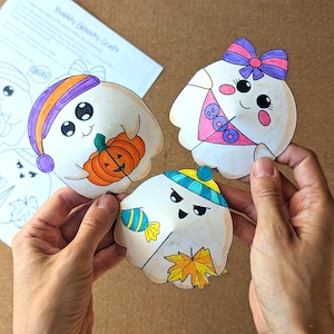 May include: Three colorful, hand-drawn ghost cutouts with various accessories, including a pumpkin, bow, and candy. The ghosts are held in a person's hands, with a sheet of paper labeled "Bubbly Ghosts Craft" in the background.