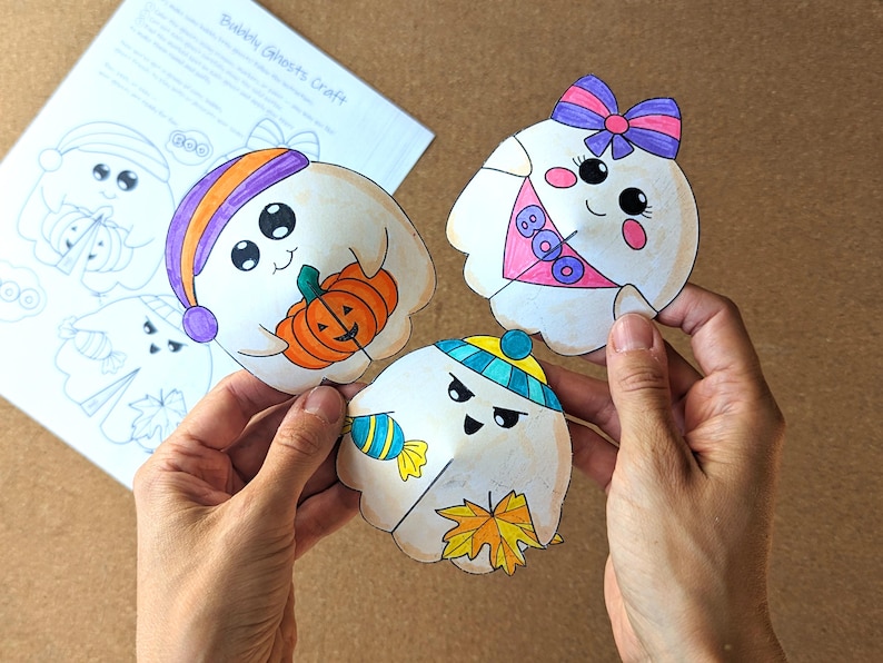 May include: Three colorful, hand-drawn ghost cutouts with various accessories, including a pumpkin, bow, and candy. The ghosts are held in a person's hands, with a sheet of paper labeled "Bubbly Ghosts Craft" in the background.