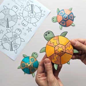 Op de afbeelding: Een knutselproject met kleurrijke, uitgesneden schildpadvormen. Een schildpad wordt door handen vastgehouden en toont een oranje en geel schildontwerp. Andere schildpadden hebben blauwe, oranje en groene schilden. Er is ook een vel papier met schildpadomtrekken aanwezig.