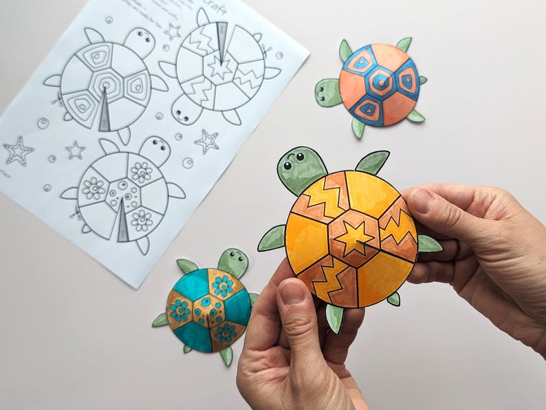 Op de afbeelding: Een knutselproject met kleurrijke, uitgesneden schildpadvormen. Een schildpad wordt door handen vastgehouden en toont een oranje en geel schildontwerp. Andere schildpadden hebben blauwe, oranje en groene schilden. Er is ook een vel papier met schildpadomtrekken aanwezig.