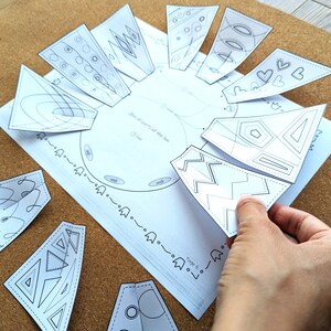 May include: A white paper craft with a circular design and several detached, triangular pieces. The pieces feature various geometric and abstract line art designs. A hand is holding one of the pieces. The craft is on a brown surface.