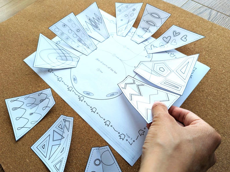 May include: A white paper craft with a circular design and several detached, triangular pieces. The pieces feature various geometric and abstract line art designs. A hand is holding one of the pieces. The craft is on a brown surface.