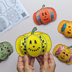 Op de afbeelding: Een set papieren pompoenknutsels met diverse kleuren en patronen, waaronder een gele pompoen met bladeren, een oranje met geometrische vormen en een groene met rode stippen, samen met een geprint instructieblad.