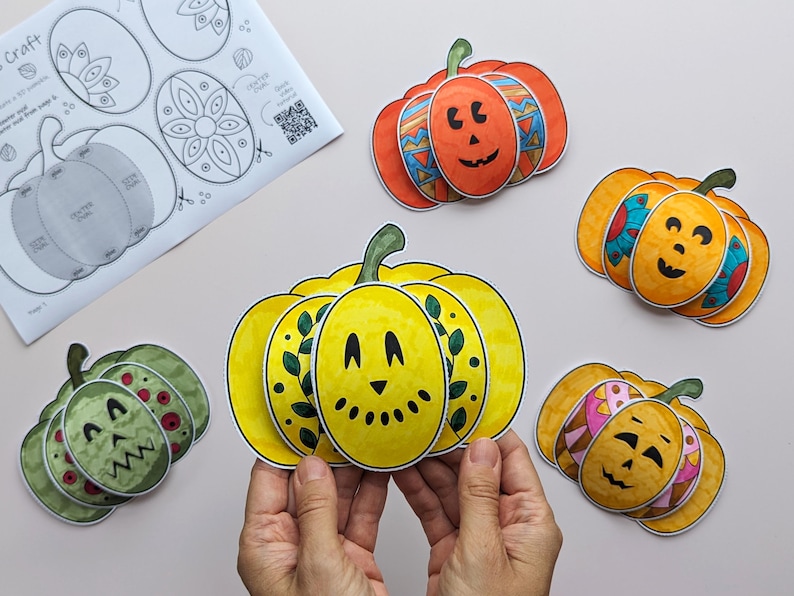 Op de afbeelding: Een set papieren pompoenknutsels met diverse kleuren en patronen, waaronder een gele pompoen met bladeren, een oranje met geometrische vormen en een groene met rode stippen, samen met een geprint instructieblad.
