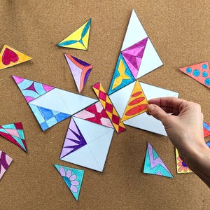 May include: A colorful butterfly puzzle made of white paper triangles with various designs. The puzzle is partially assembled, with a hand placing a triangle. The triangles feature patterns in pink, blue, yellow, and orange. The background is a brown corkboard.