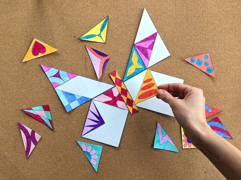 May include: A colorful butterfly puzzle made of white paper triangles with various designs. The puzzle is partially assembled, with a hand placing a triangle. The triangles feature patterns in pink, blue, yellow, and orange. The background is a brown corkboard.