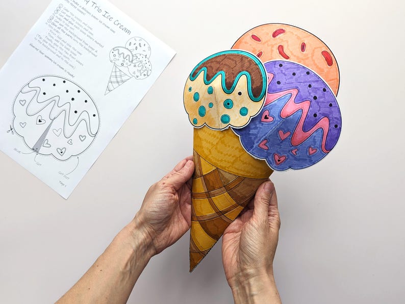 Op de afbeelding: Een handgemaakte papieren ijshoorntje met drie bollen in bruin, oranje en paars. Het hoorntje wordt door handen vastgehouden. Een zwart-wit tekening van het ijs is zichtbaar op de achtergrond. De tekst "My Trio Ice Cream" is zichtbaar.
