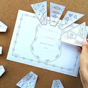 May include: A white paper with a diagram of a foot, surrounded by several white, geometric cutouts with black line art. The text "Glue all parts of the foot" is visible. The cutouts are being held by a hand.