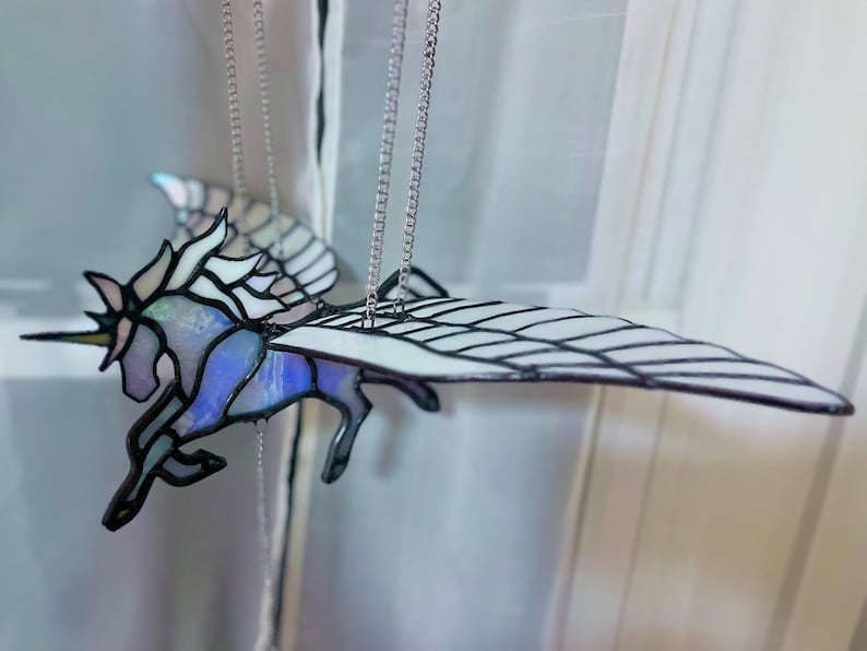May include: A stained glass winged unicorn mobile with iridescent glass and black metal accents. The unicorn is suspended from a silver chain.