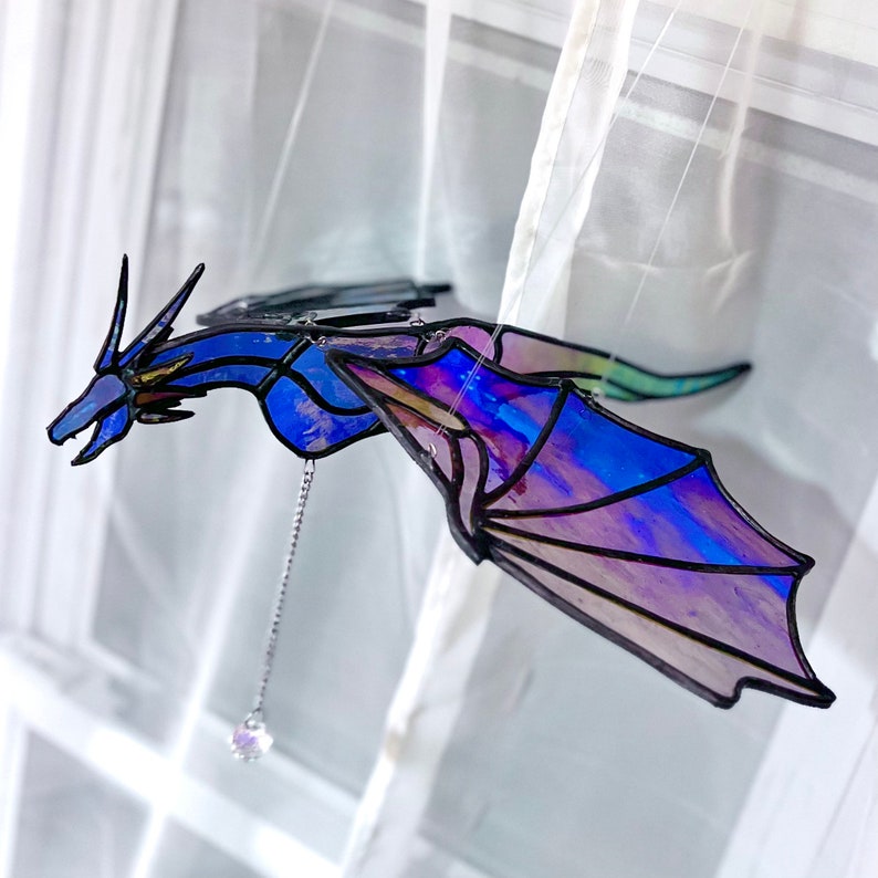 Puede incluir: Un m&oacute;vil de vidrio coloreado con un drag&oacute;n con alas iridiscentes azules y moradas. El drag&oacute;n est&aacute; suspendido de una cadena con un cristal colgando en la parte inferior.