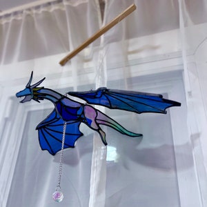 Puede incluir: Un m&oacute;vil de vidrio coloreado con un drag&oacute;n con alas iridiscentes azules y moradas, colgando de un techo blanco con un prisma de cristal en el extremo.