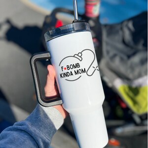 May include: White stainless steel tumbler with a black handle and a red star. The tumbler has a black and white graphic that says "F-Bomb Kinda Mom" with a stylized image of a baby bottle.