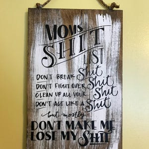May include: A wooden sign with a rope hanger. The sign has a distressed white and brown background with black lettering that reads "Moms Shit List". The list includes rules like "Don't Break Shit" and "Don't Make Me Lose My Shit".