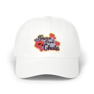 Può includere: Cappellino da baseball bianco con visiera curva. Il cappellino presenta una toppa ricamata con le parole "Toxica Pero Chula" in viola, giallo e rosa, insieme a un design di labbra rosse e accenti a cuore.