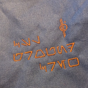 May include: Orange embroidered symbols on a blue fabric background. The symbols include a series of triangles, squares, and lines. The text "17701" is also embroidered in orange.