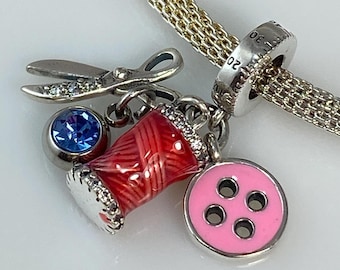 May include: A silver charm bracelet with a red and white spool of thread, a pink button with four holes, a blue rhinestone, and a pair of silver scissors.