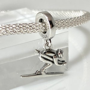 May include: Silver charm featuring a skier in a downhill position on skis. The charm is attached to a silver chain bracelet.