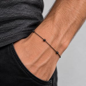 May include: A delicate black chain bracelet featuring three evenly spaced, small, smooth black spherical beads. The bracelet is constructed from thin, dark metal links that form a continuous, minimalist loop around the wrist area.
