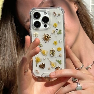 Peut inclure: Coque de téléphone transparente ornée de fleurs séchées, d'éléments dorés et d'un motif en forme d'éclat de soleil. L'étui présente une variété de petites fleurs pressées en jaune, blanc et vert, créant un aspect naturel et artistique. La coque est tenue par une personne.