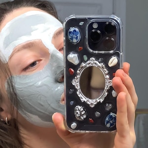 May include: A clear phone case with a black interior, adorned with decorative elements including shells, gemstones, and ceramic pieces. The case features a silver-framed mirror and a clear protective border.
