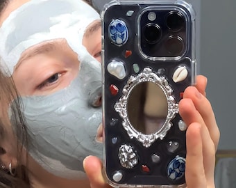 May include: A clear phone case with a black interior, adorned with decorative elements including shells, gemstones, and ceramic pieces. The case features a silver-framed mirror and a clear protective border.