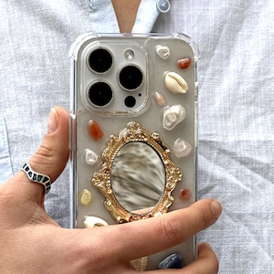 May include: Clear phone case with embedded seashells, gemstones, and a gold-framed mirror. The case is held in a hand, showcasing the decorative elements and the phone's camera lenses.
