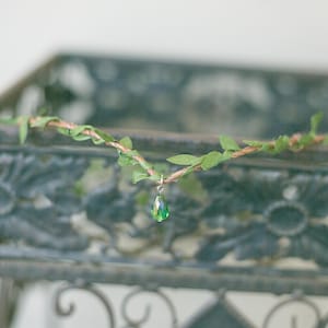 May include: A green crystal pendant hangs from a delicate silver chain, adorned with a leafy vine made of green fabric. The vine is draped over a black metal frame.