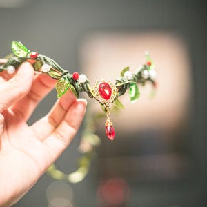 Peut inclure: Une couronne de feuilles vertes et dorées avec des pierres précieuses rouges et des perles blanches. La couronne a un pendentif en pierre précieuse rouge.