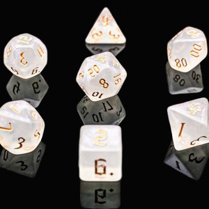 Op de afbeelding: Een set van zeven witte dobbelstenen met gouden nummers. De dobbelstenen liggen op een zwart oppervlak en reflecteren licht.