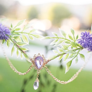 May include: A floral crown featuring purple flowers, green leaves, and a gold-colored metal centerpiece with a teardrop-shaped purple gem and dangling crystal. The crown is designed for special occasions.