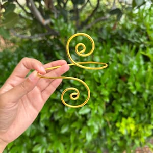May include: A gold-colored arm cuff with a spiral design. The cuff is held in front of a green leafy background. The cuff has a simple, elegant design, with a curved shape that wraps around the arm.