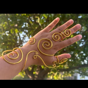 May include: Gold-coloured wire hand jewellery featuring spiral and swirl designs. The jewellery includes a bracelet, rings, and decorative elements on the back of the hand. The design is artistic and unique.
