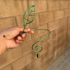 May include: A green wire arm cuff featuring a leaf design and a dangling green gem. The cuff has a spiral design and is held up by a hand. The leaf is made of wire and has a detailed vein pattern.