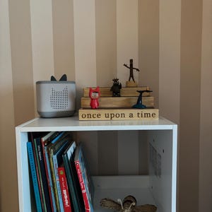 Pu&ograve; includere: Una mensola bianca con libri e oggetti decorativi. Un altoparlante grigio con orecchie &egrave; accanto a un cartello di legno che recita "once upon a time". Sono esposte anche varie statuette e una falena tessuta.