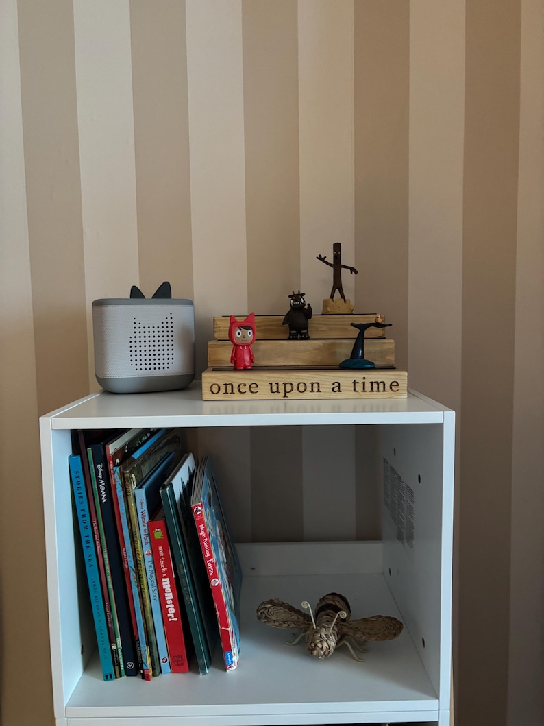 Pu&ograve; includere: Una mensola bianca con libri e oggetti decorativi. Un altoparlante grigio con orecchie &egrave; accanto a un cartello di legno che recita "once upon a time". Sono esposte anche varie statuette e una falena tessuta.