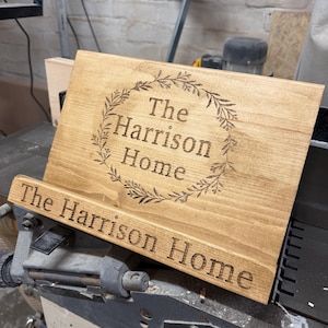 May include: A light brown wooden sign engraved with the words "The Harrison Home". The sign features a decorative wreath around the text and is also engraved on the base. The sign is made of wood.