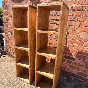 May include: Two tall, narrow wooden shelving units with four open compartments each. The left unit features the engraved text "The Warmth Of The Sun" on the top edge, while the right unit features the engraved text "Sail On Sailor" on the top edge.