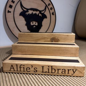 Pu&ograve; includere: Un espositore in legno a tre livelli con dettagli neri e la scritta incisa "Alfie's Library". Un cartello circolare in legno con "The Rolling Cow" e una silhouette di mucca delle Highlands &egrave; appeso sullo sfondo. L'espositore &egrave; realizzato in legno chiaro.