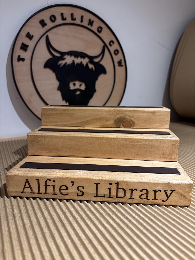 Pu&ograve; includere: Un espositore in legno a tre livelli con dettagli neri e la scritta incisa "Alfie's Library". Un cartello circolare in legno con "The Rolling Cow" e una silhouette di mucca delle Highlands &egrave; appeso sullo sfondo. L'espositore &egrave; realizzato in legno chiaro.