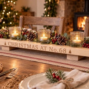 May include: A rectangular light wood centrepiece featuring the engraved text CHRISTMAS AT No.68 flanked by two small tree icons. It holds three ribbed glass votive candle holders surrounded by pine branches, pinecones, and red berries.