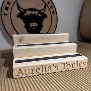 Pu&ograve; includere: Un espositore in legno a tre livelli con dettagli neri e la scritta incisa "Aurelia's Tonies". Un cartello in legno con "THE ROLLING COW" e una silhouette di mucca delle Highlands sullo sfondo.